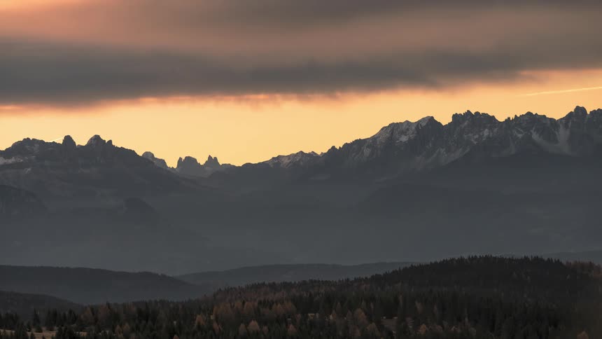 A dark wall cloud hangs over a vast mountain range rolling hills and a forest