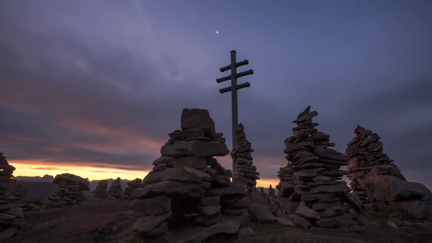 A wooden cross between several piles of small stones and the sky is with several clouds and it is dawn
