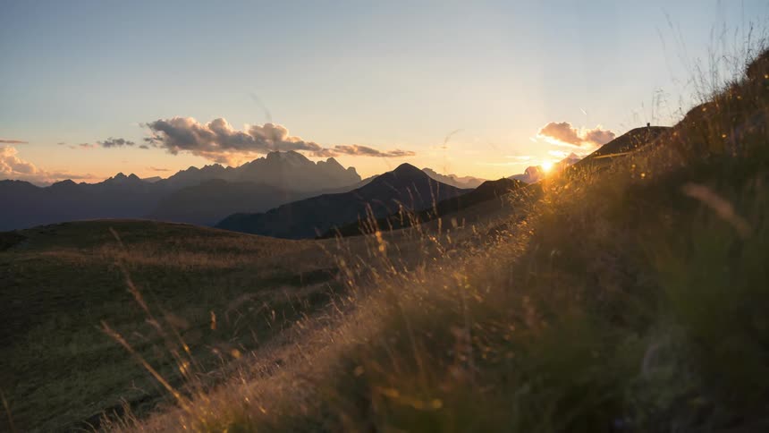 A field with some hills covered in grass and the sky with clouds passing by and the sun hiding behind mountains