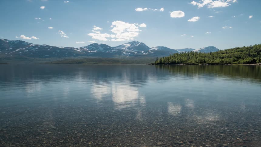 The still lake reflects floating clouds and the snow capped peaks of the mountains