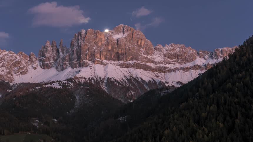 Mountainous landscape with snowcapped peaks and the moon rising on the horizon as dusk falls