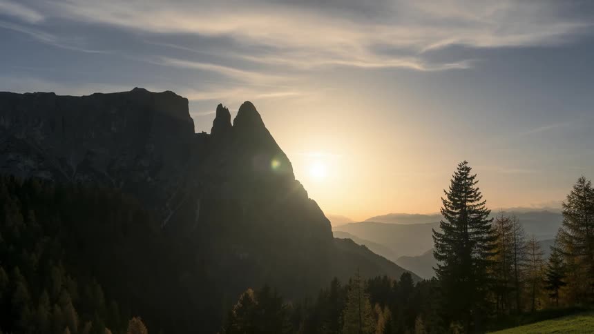 Sun peeking out from behind tall mountains behind a forest
