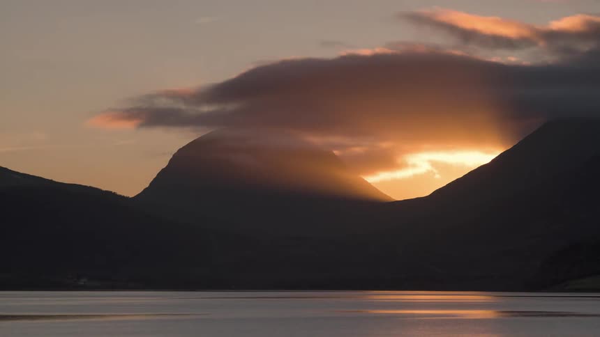 Sunset with orange tones visible through an opening between dark mountains and a large body of water