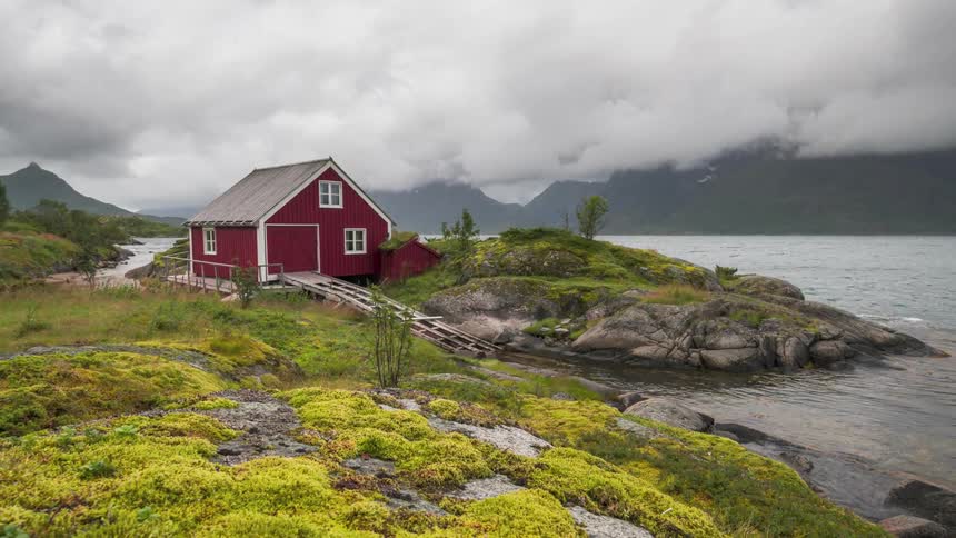 Red wooden house by a lake surrounded by mountains on a cloudy day