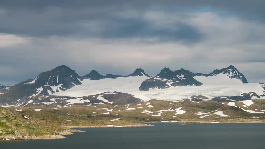 Snowy mountains near a beach with sand and grass under fastmoving clouds