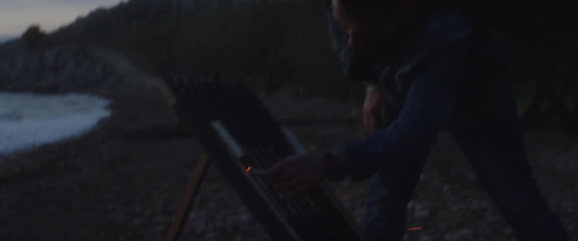 A man lighting fireworks with the burning end of his cigarette next to a body of water