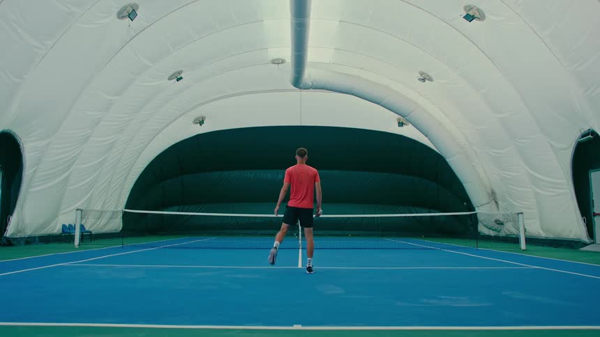 Man enters a bubble tennis court and begins warming up while facing the net