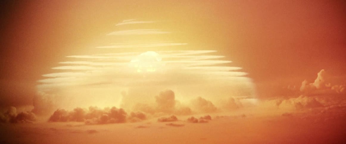 An atomic bomb detonates in the distance creating a large mushroom and shockwaves