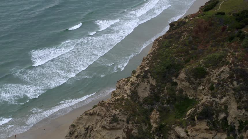 Aerial view of a beach with gentle waves on the sand and a lush green cliff