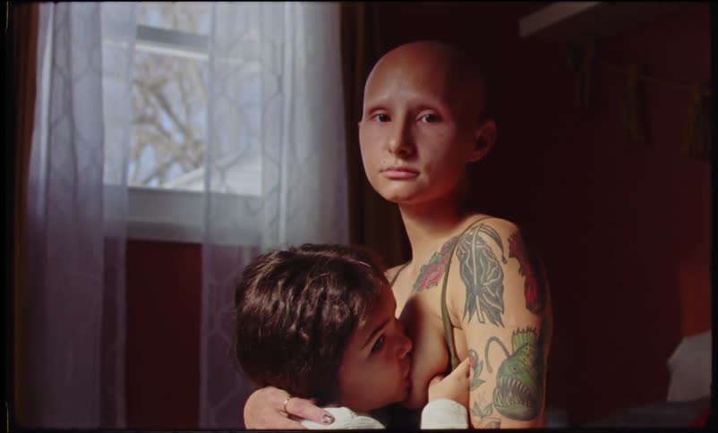 A bald white woman breastfeeds her child in a bedroom