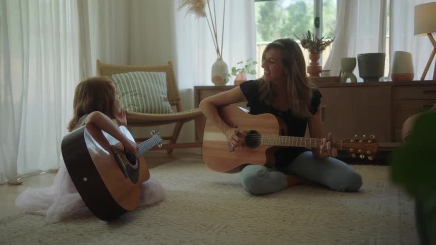 A mother and daughter play acoustic guitars together in a living room