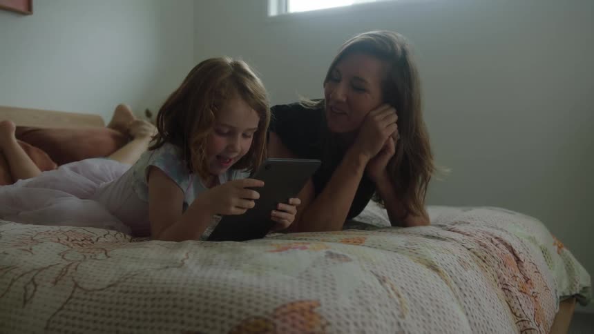 A mother and her daughter who is playing on her cell phone are lying on a bed and they give each other a loving kiss on the cheek and are happy