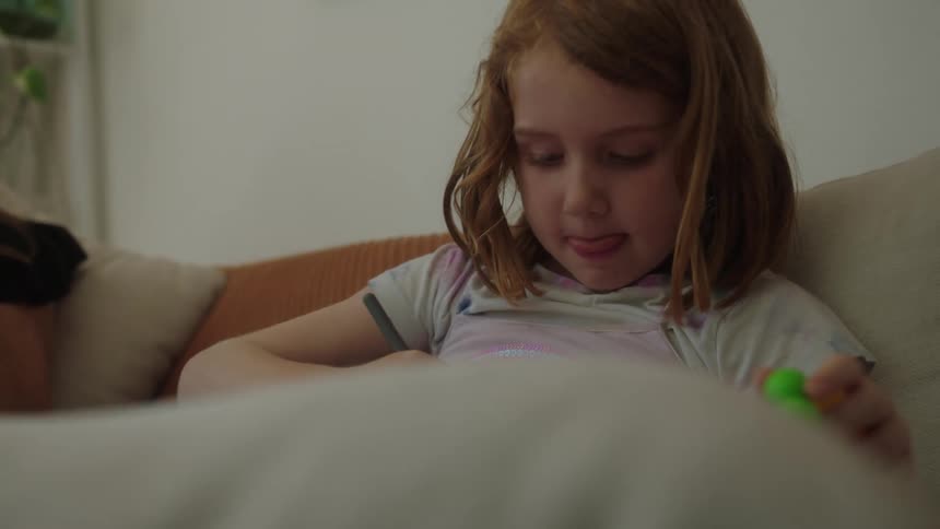 A mother on a smartphone smiles at her daughter who is drawing while sitting on a couch