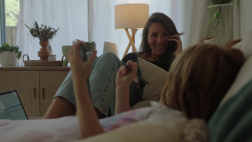 Mother talking on the phone while her daughter draws on a tablet both are sitting on a sofa in the living room