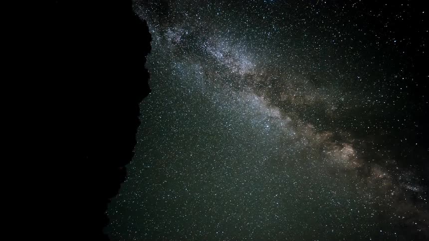 Clouds move across the milky way seen in a night sky and disappear behind a mountain