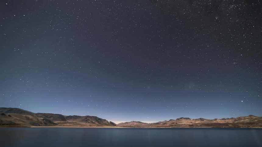 Starry night sky with the milky way that dawns with a blue sky and stretches across a mountainous horizon on the edge of a lake