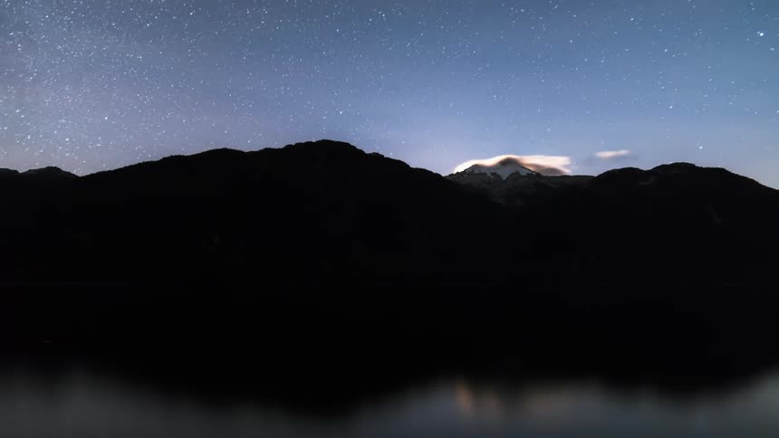 Landscape of a dusk sky with the aurora borealis appearing above a mountain on the edge of a lake