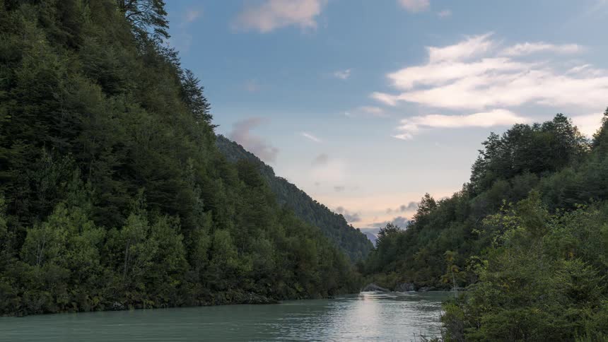 Clouds flow over stream passing forested mountains as the sun sets