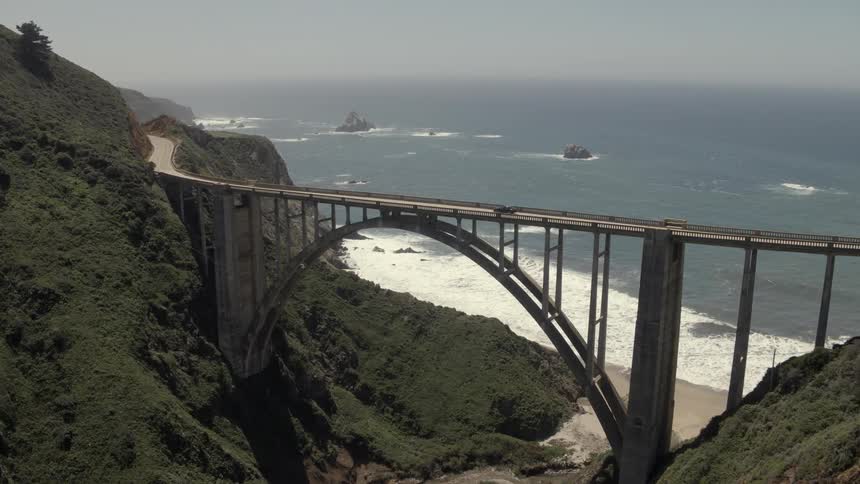 Cars drive over a tall bridge with the white ocean waves in the background