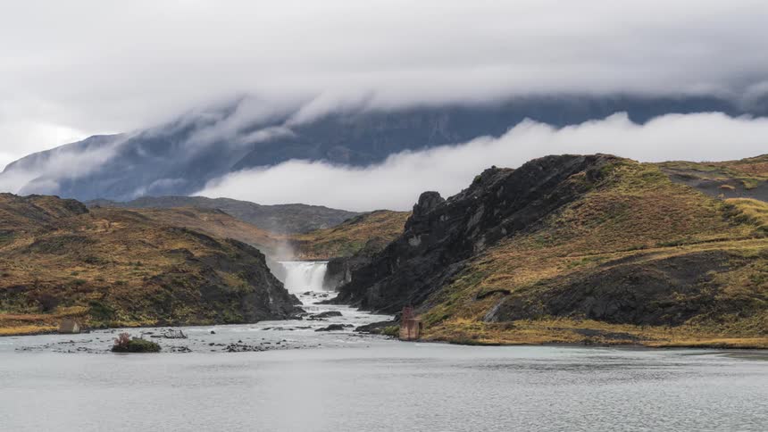 Scenery of a waterfall surrounded by mountains by a lake and low clouds