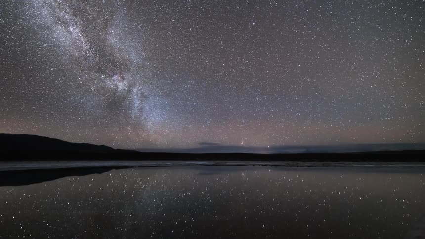 A landscape of a lake reflecting the sky at night and with many stars and time is passing quickly