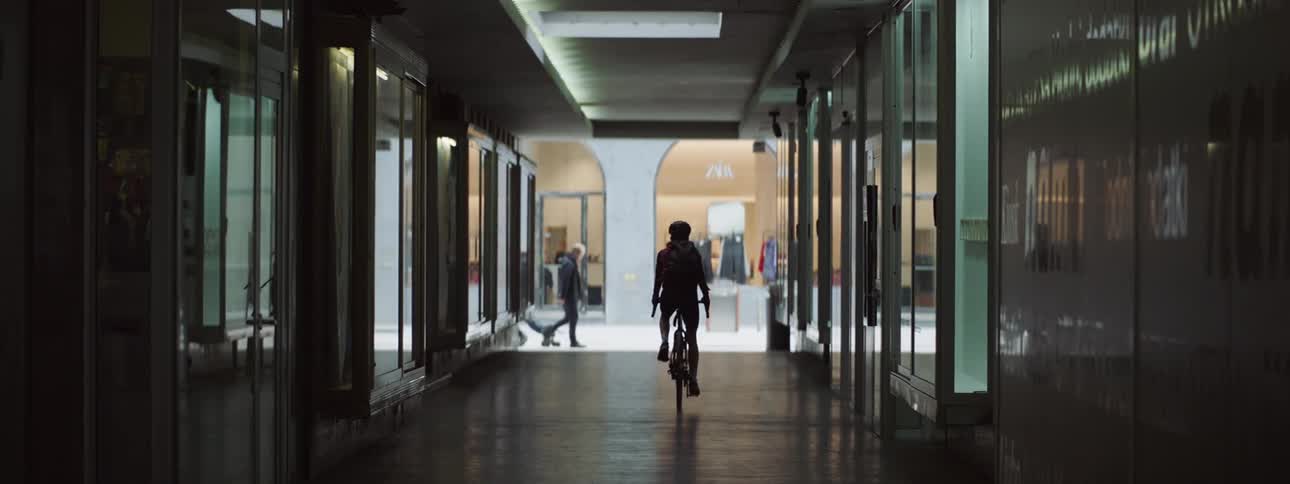 Cyclist rides through dark corridor turning onto bright area at busy shopping mall