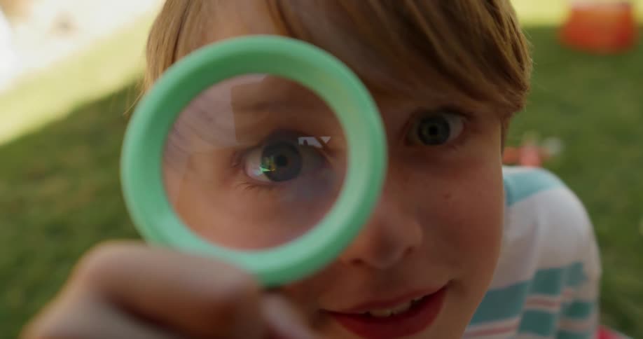 Child holds a magnifying glass in front of their eye while sitting in a grassy yard
