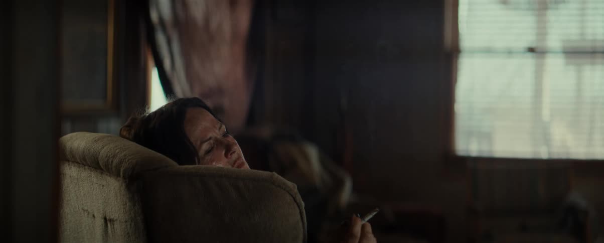 A woman resting her head on a sofa armrest while smoking in a dimly lit room