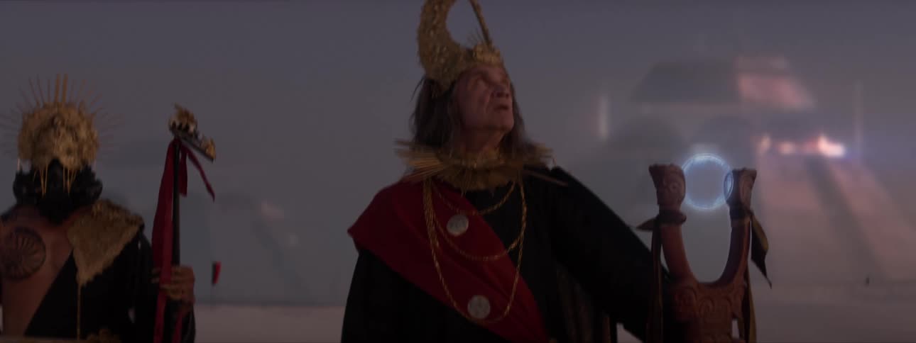 Two people in mythological costumes with one of them holding a staff and pyramids in the background