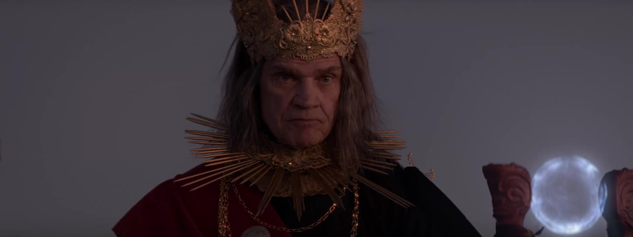 A man dressed like a wizard angrily speaks while a blue orb shines next to him