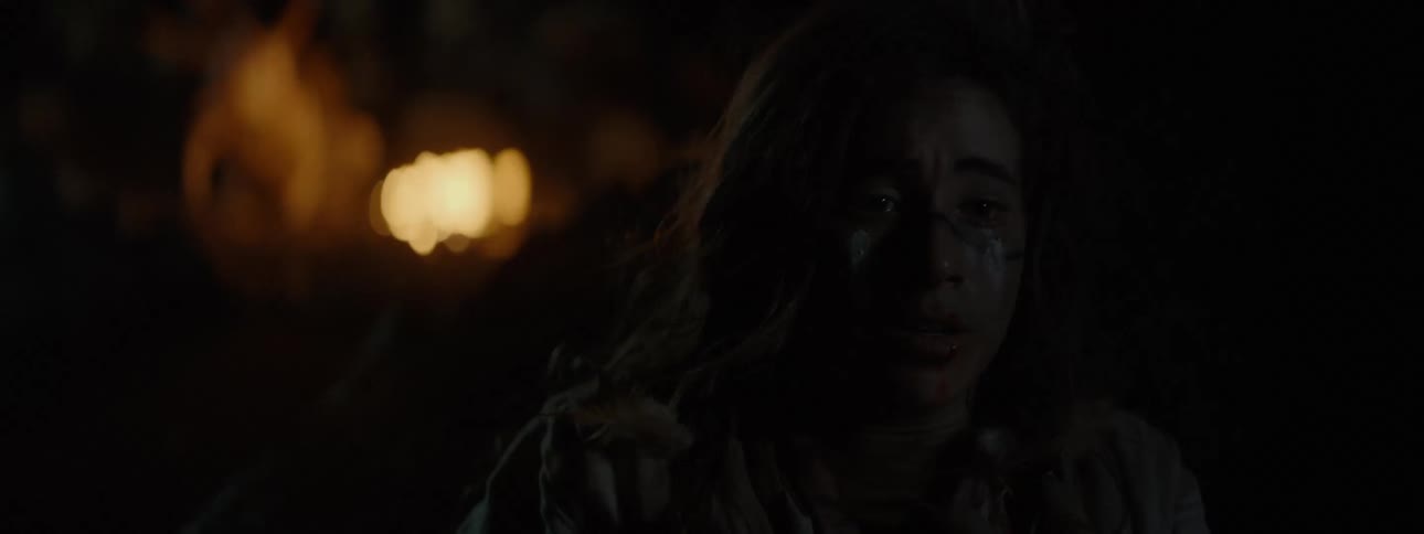 A woman is distressed and scared behind a rock and she looks at a bonfire that is behind her and quickly hides