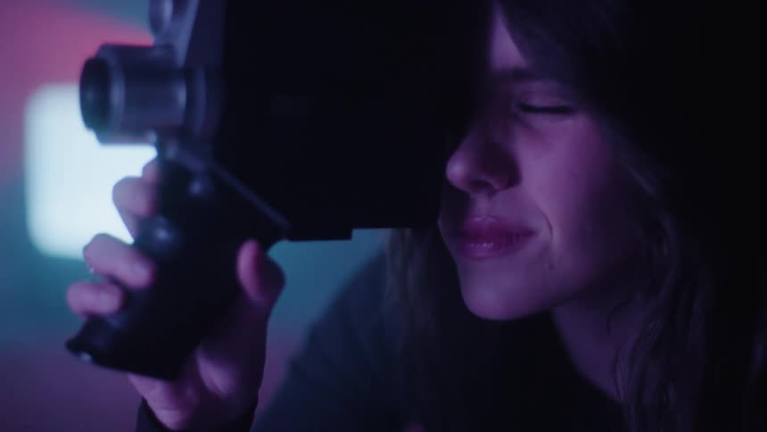 A closeup of a woman holding a vintage film camera with a background of purple and red hues