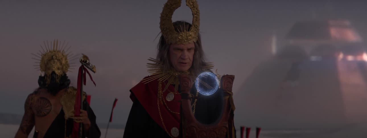 Two men in costumes in a desert environment with one of them holding a magical circular staff and futuristic pyramids in the background