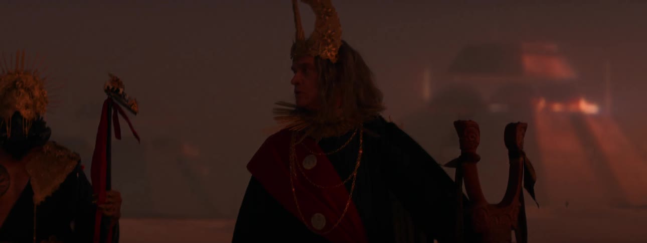 A man wearing a crown standing in front of futuristic pyramids looks around when the lights change