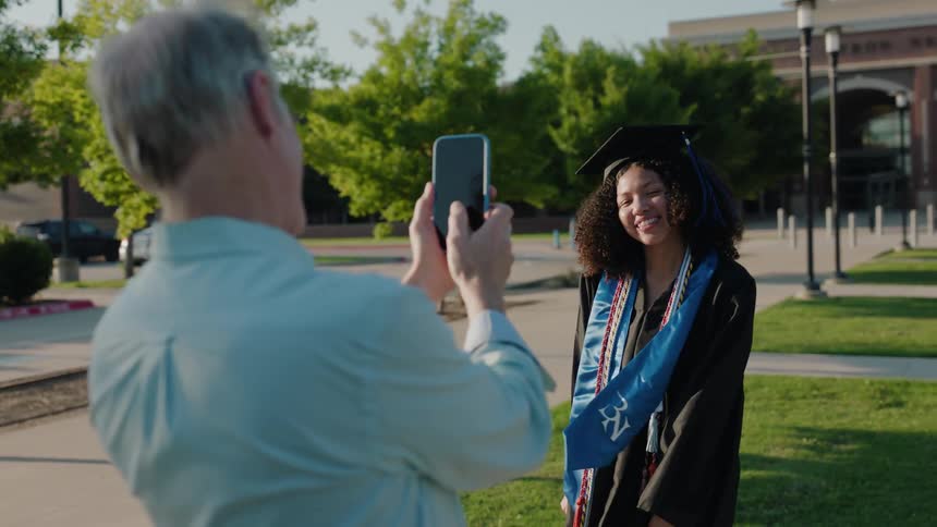 Father taking a photo with a smartphone of his daughter who is smiling and posing in a graduation gown
