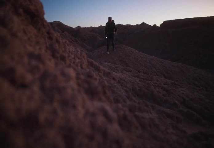 Man with a backpack and holding a professional camera walking on rocky terrain during twilight