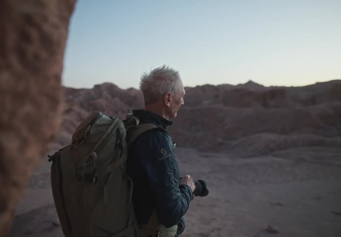 Man with gray hair with a backpack looking at a desert terrain with rock formations at dusk and holding a professional camera