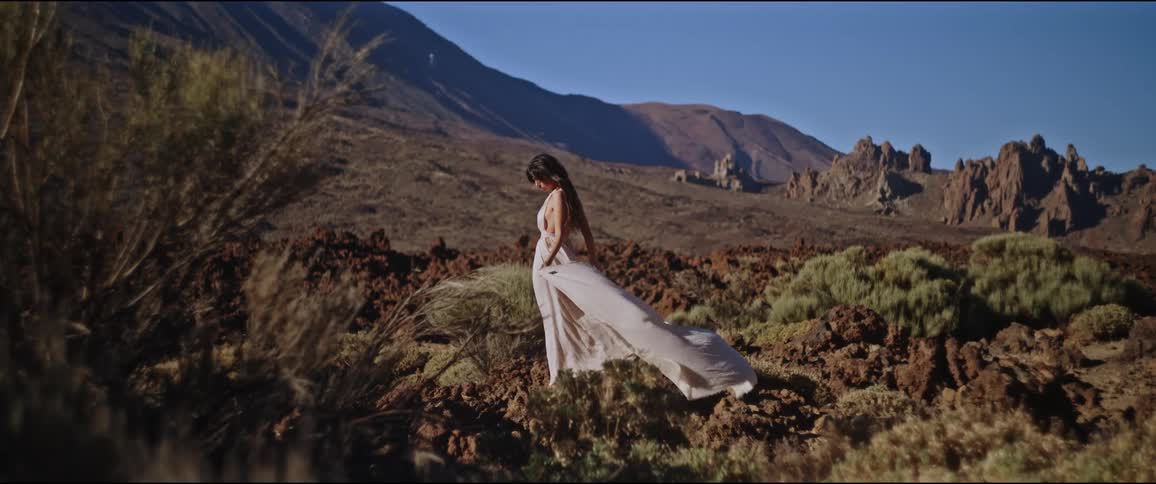 Woman in a long white dress standing on rocky terrain with sparse vegetation