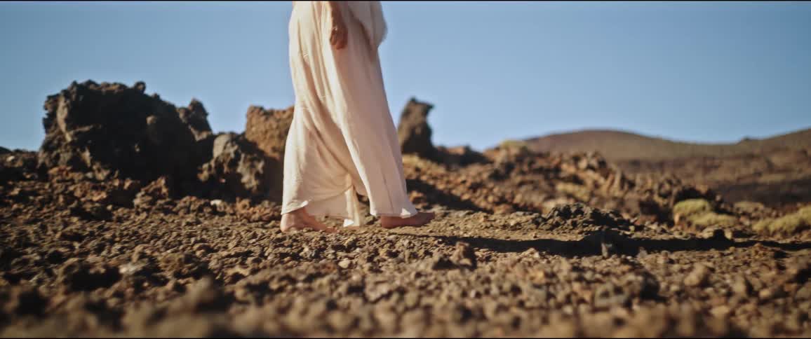Woman wearing a long white dress and barefoot walking along the rocks with mountains around
