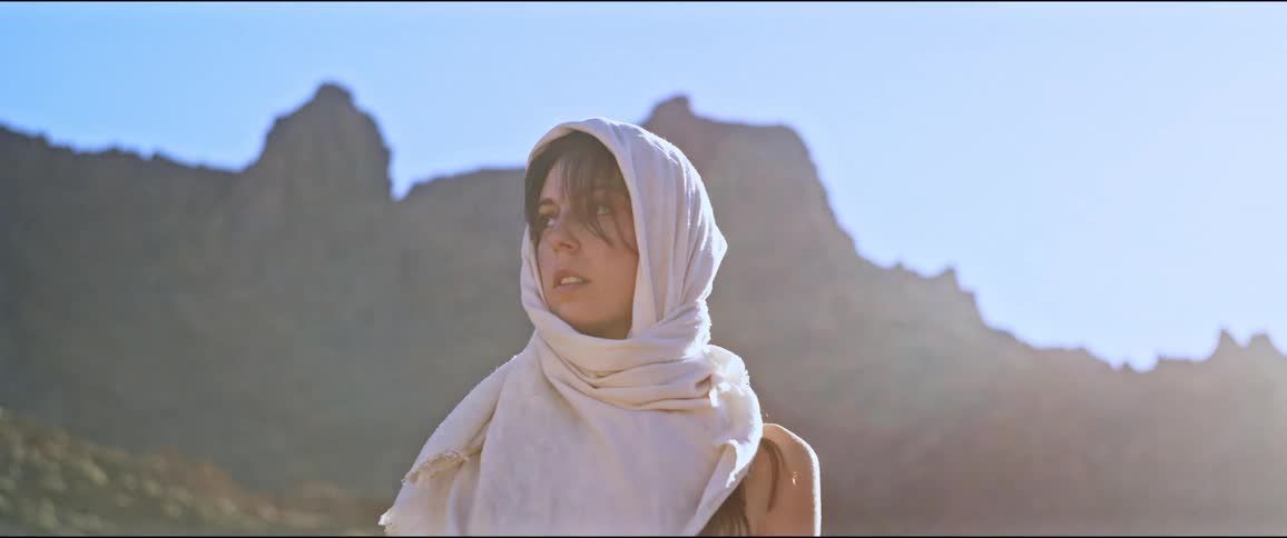 A white woman wearing a head scarf looks around her nervously against a mountain backdrop