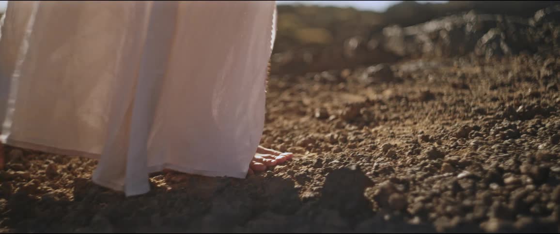 The legs of a barefoot person walking on a stone floor and she is wearing a long white dress