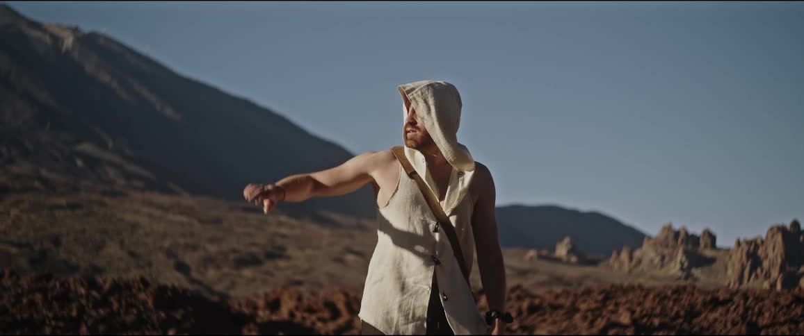 Man in a hooded sweatshirt raising his hand against the sky in a natural environment surrounded by rocks and mountains