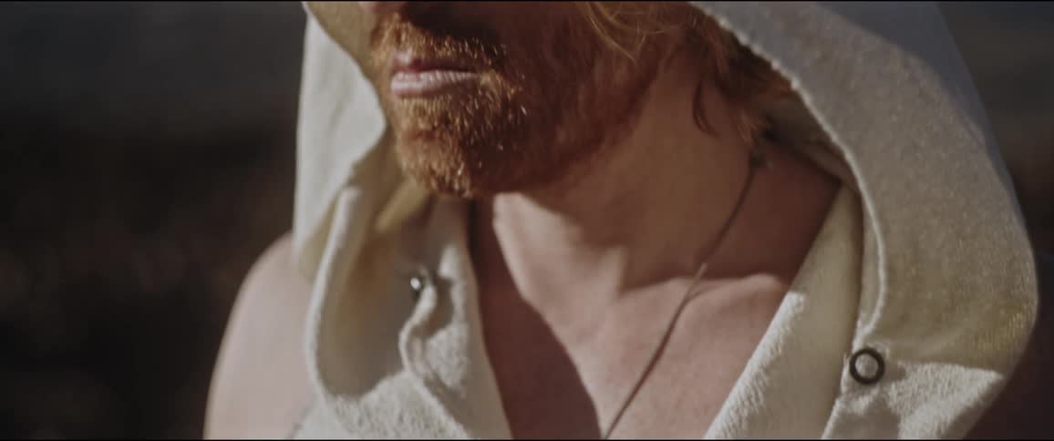 A bearded man with long hair wearing a sleeveless hoodie in nature