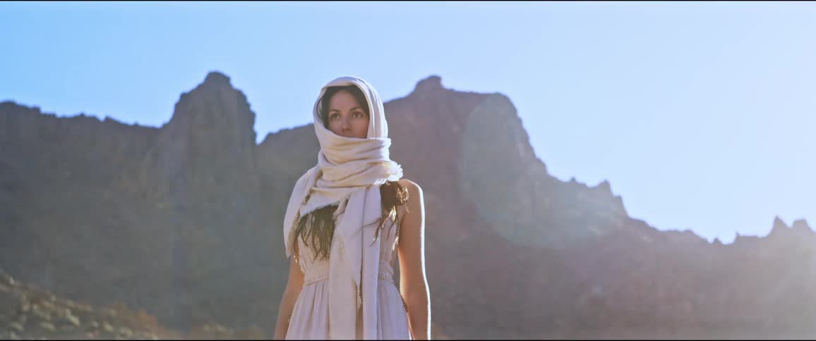 A woman wearing a scarf over her head stands in front of a mountain under a blue sky in a sunny day