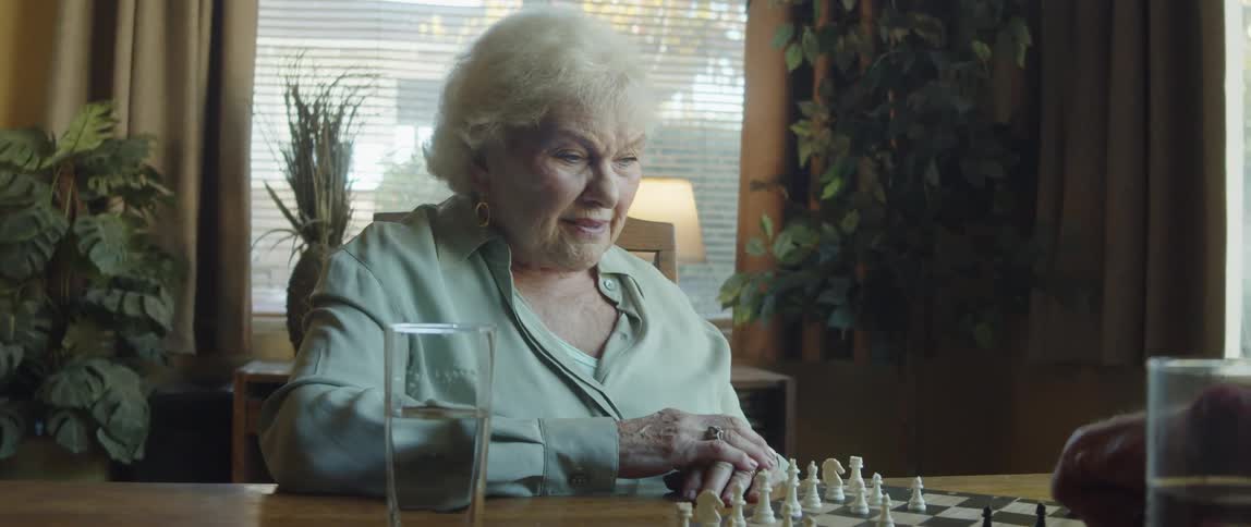 An elderly man and woman are playing chess on a wooden table and talking