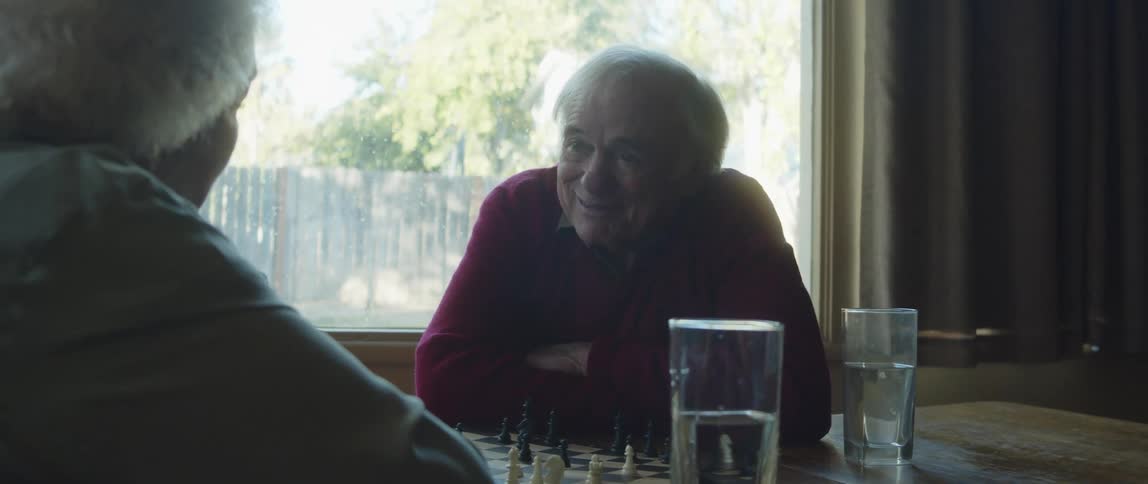 Two elderly people sitting at a table with a chess set talking