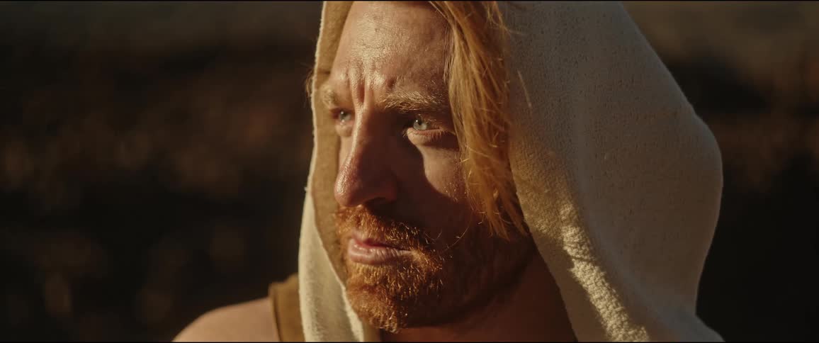 A bearded man with long hair wearing a sleeveless hoodie in nature