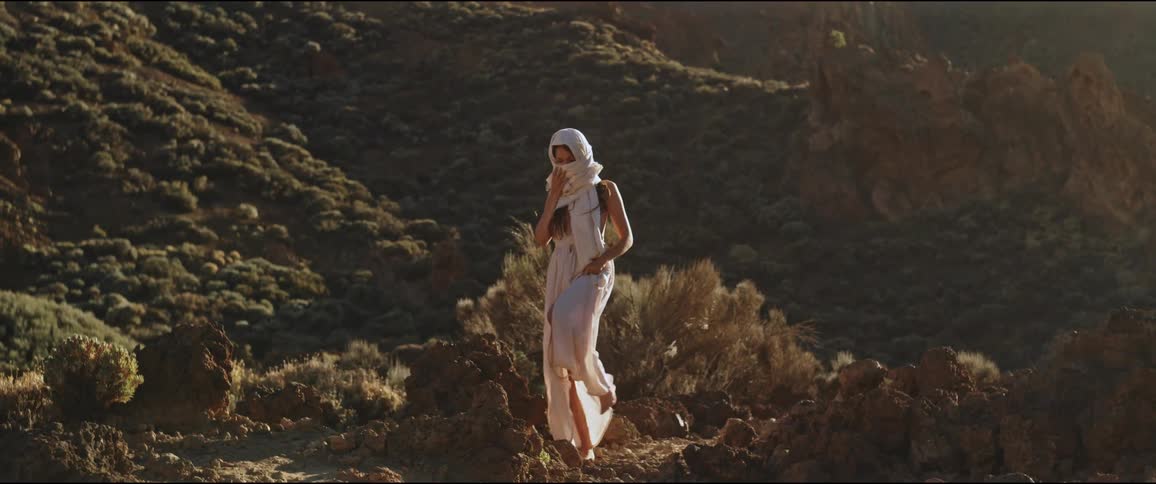 Woman in a white dress and headscarf walking alone through arid rocky terrain