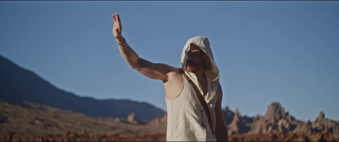 A bearded man with long hair wearing a sleeveless hoodie holds a hand up in front of a mountain