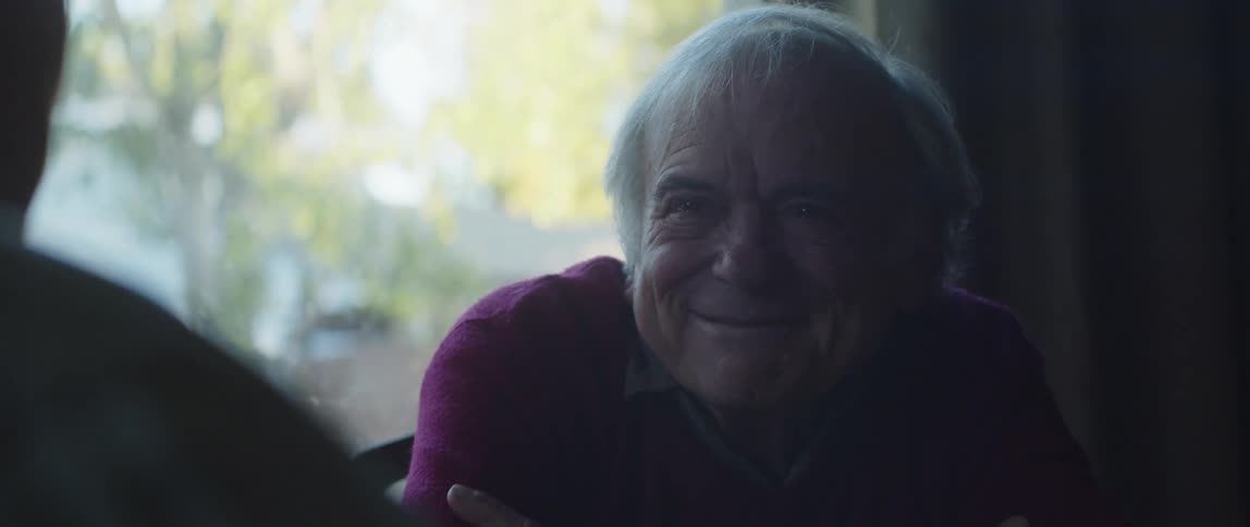 An elderly man laughs and enjoys conversing with another person in his house